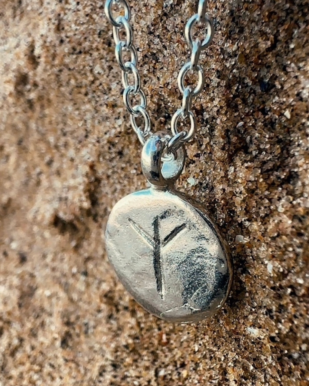 Close-up of a sterling silver Algiz rune charm showing hand-etched detailing and a worn, relic-inspired texture on sand.