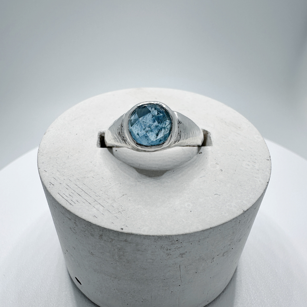 Front view of a sterling silver aquamarine signet ring with natural inclusions displayed in a contemporary concrete ring holder.