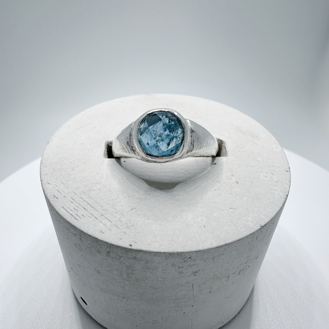 Front view of a sterling silver aquamarine signet ring with natural inclusions displayed in a contemporary concrete ring holder.