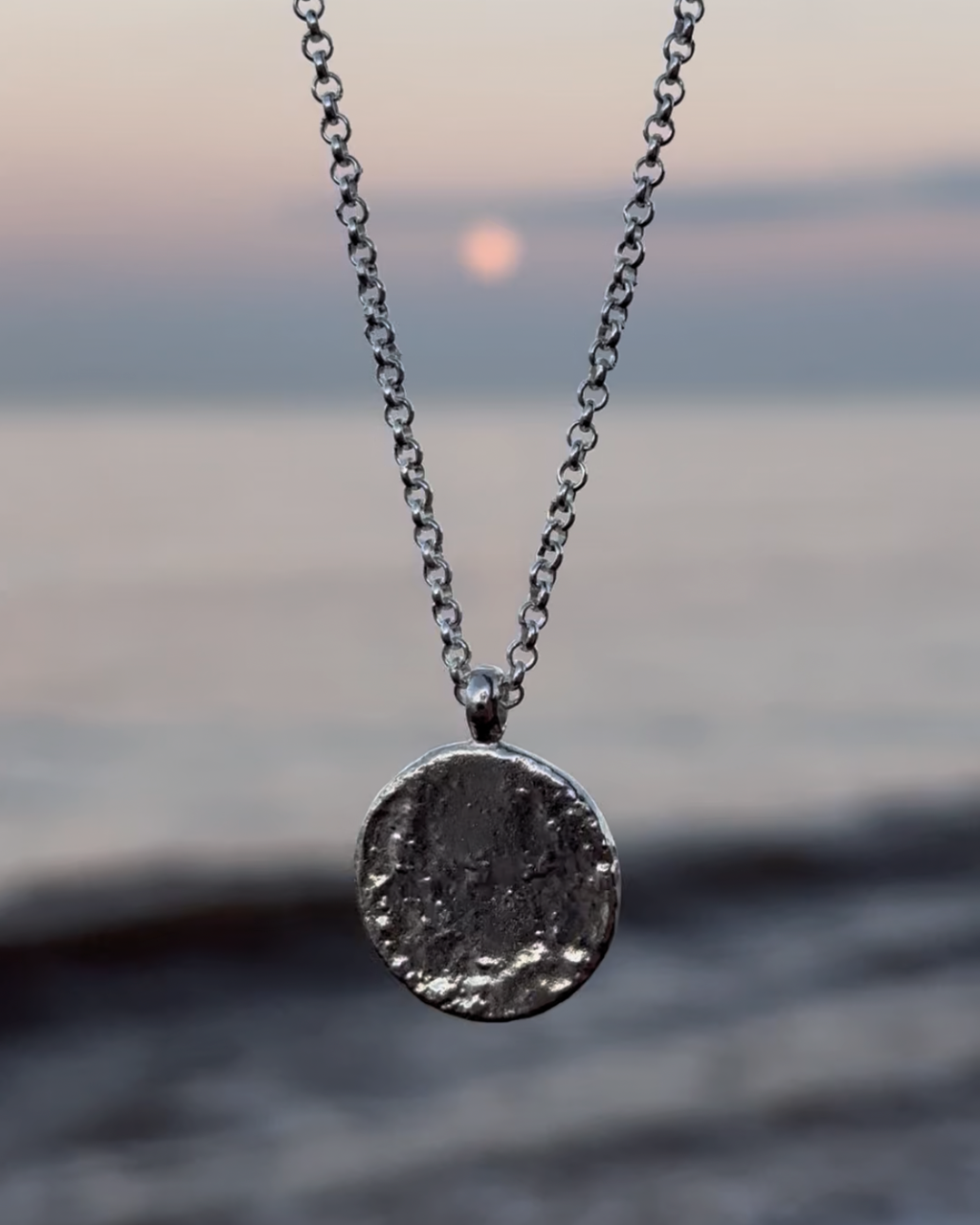 Sterling silver full moon necklace hanging in front of a blurred moonrise, sea background with purple, blue and pink sunset colours.