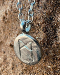 Close-up of a sterling silver Mannaz rune charm with hand-etched detailing and a relic-like texture on a sand background.