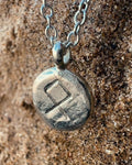 Close-up of a sterling silver Othila rune charm showcasing hand-etched lines and a relic-style textured surface on sand.