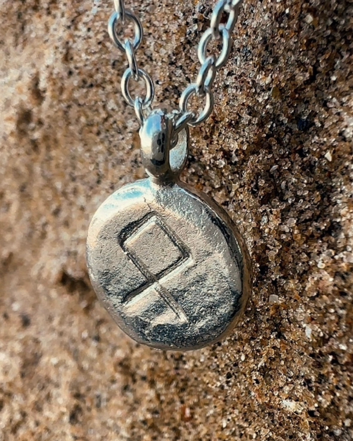 Close-up of a sterling silver Othila rune charm showcasing hand-etched lines and a relic-style textured surface on sand.