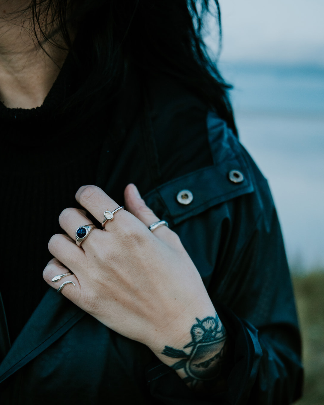 Sterling silver snake ring worn on the pinky finger with black clothing and a blurred coastal backdrop.
