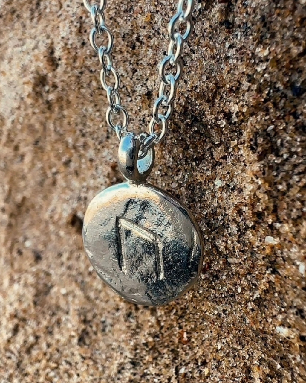 Close-up of a sterling silver Uruz rune charm with carved rune detail and a rugged, relic-like texture on a sand background.