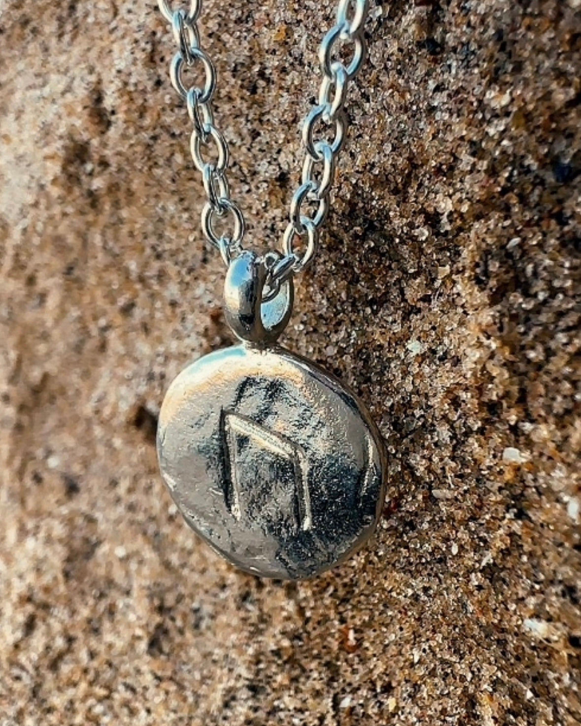 Close-up of a sterling silver Uruz rune charm with carved rune detail and a rugged, relic-like texture on a sand background.
