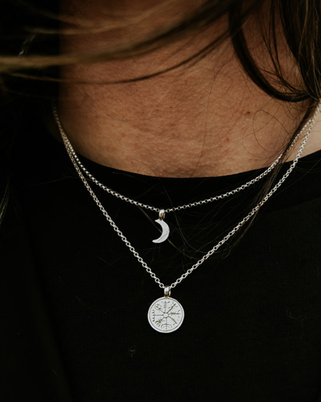 Close-up of a sterling silver Vegvisir necklace layered with a small crescent moon necklace worn with black clothing.
