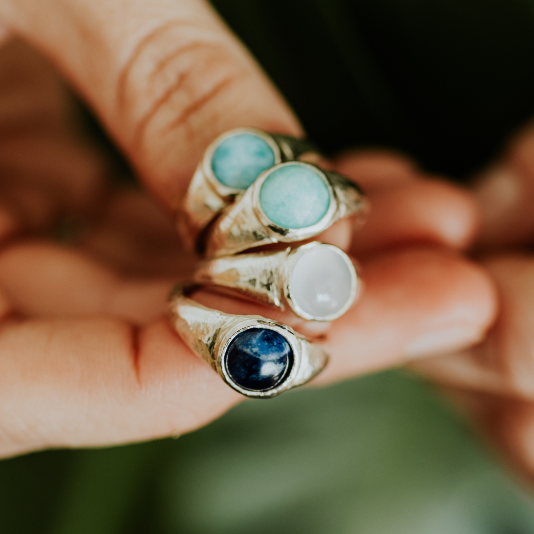 Close-up of four gemstone rings held between fingers, featuring larimar, amazonite, chalcedony, and sodalite stones.